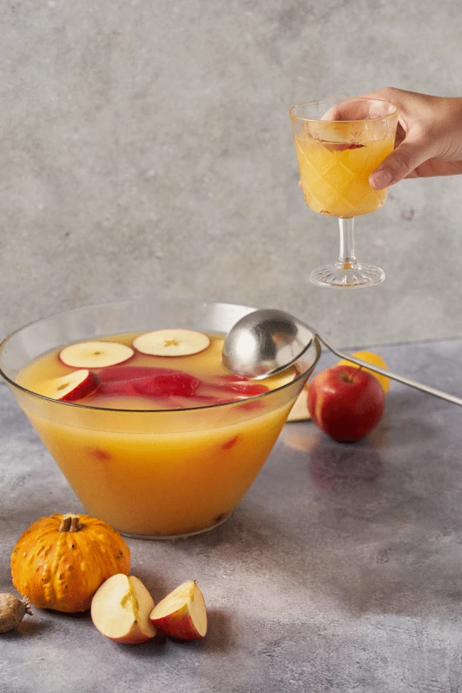 Hand hält ein Glas Apfelpunsch über einer Schüssel, umgeben von Äpfeln und einem Kürbis auf grauer Oberfläche.