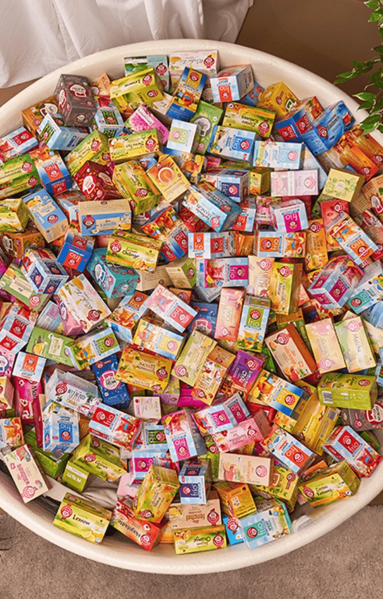 Weiße Teetasse, die mit bunten Teepackungen überquillt, auf einer braunen Fläche mit grünen Pflanzen und Stoff.