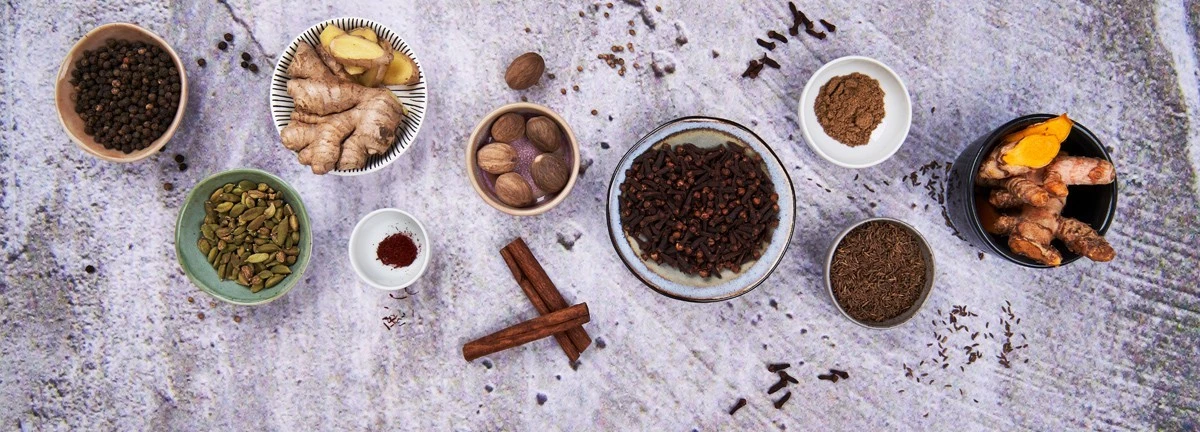 Verschiedene ganze Gewürze und Kräuter in Schalen auf einer strukturierten grauen Oberfläche, darunter Safran und Zimt.