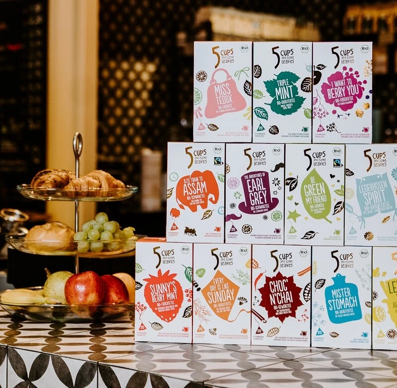 Pyramidenförmig gestapelte bunte Teeboxen auf der Café-Theke; im Hintergrund Gebäck, Obst und Barista.