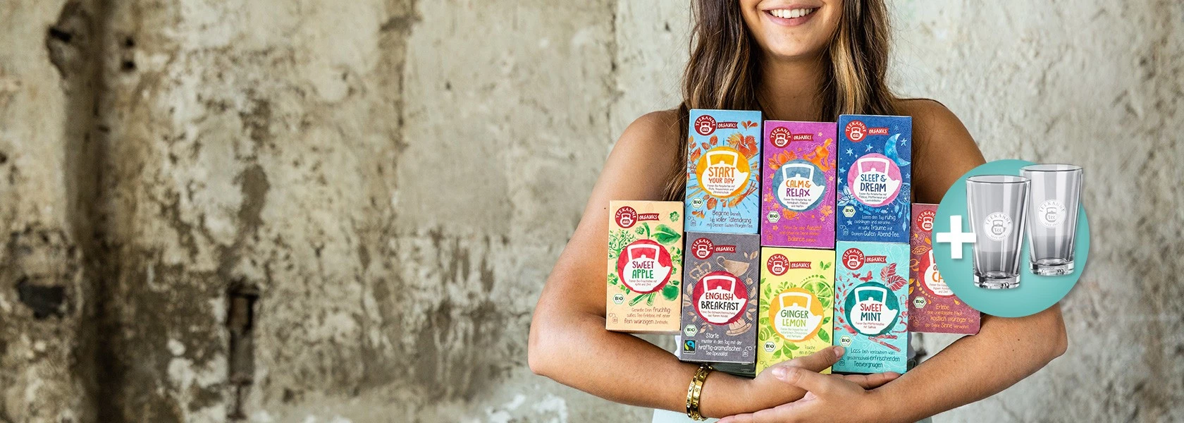 Lächelnde Frau, die bunte Teedosen mit Trinkgläsern neben sich vor einem rustikalen Wandhintergrund hält.