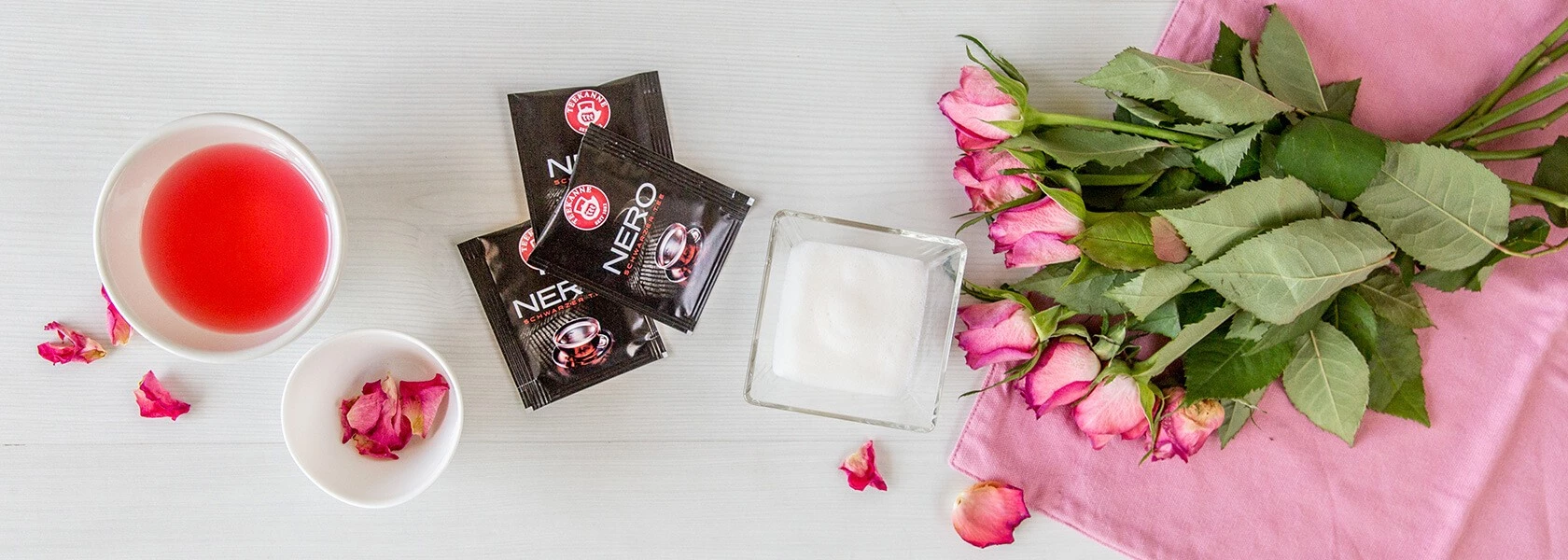 Schüssel mit roter Flüssigkeit, getrockneten Rosenblättern, Kaffeebeuteln, Zuckerdose und rosa Rosen.