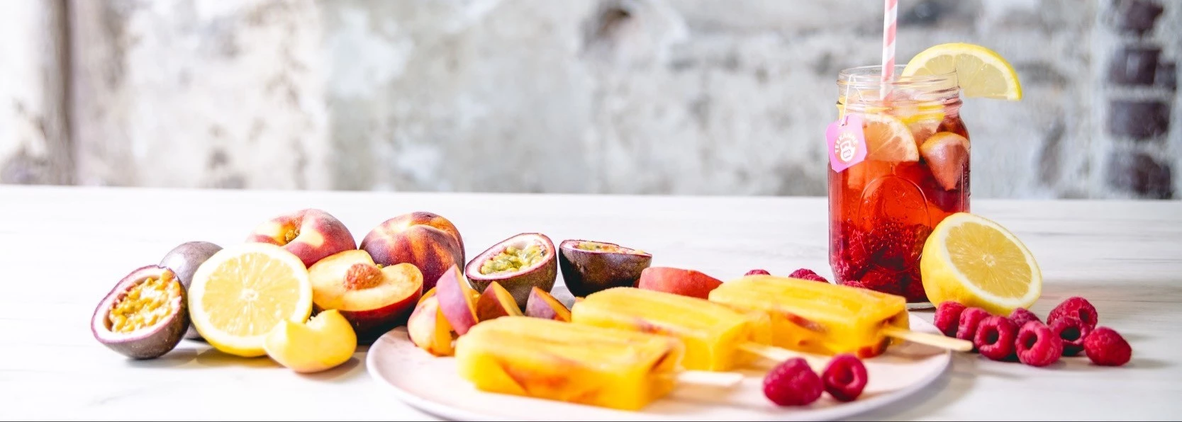 Gelbe Fruchteis am Stiel mit frischen Früchten und Eistee auf einem weißen Tisch.