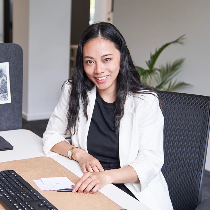 Lächelnde Frau mit langem dunklem Haar schreibt an einem Schreibtisch, Tastatur und Pflanze im Hintergrund.