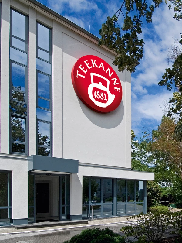 Modernes Gebäude mit großen Fenstern und TEEKANNE 1882-Teekannenschild, umgeben von Bäumen.