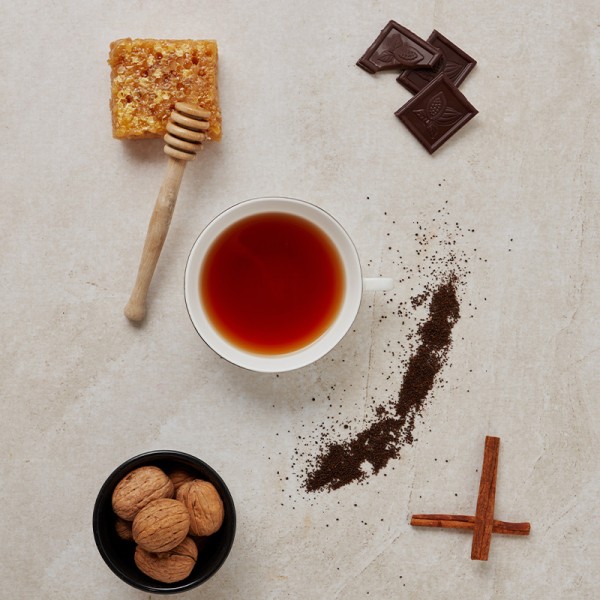 Tasse Tee mit Honigwabe, Schokolade, Walnüssen, losen Teeblättern und Zimtstangen auf hellem Hintergrund.
