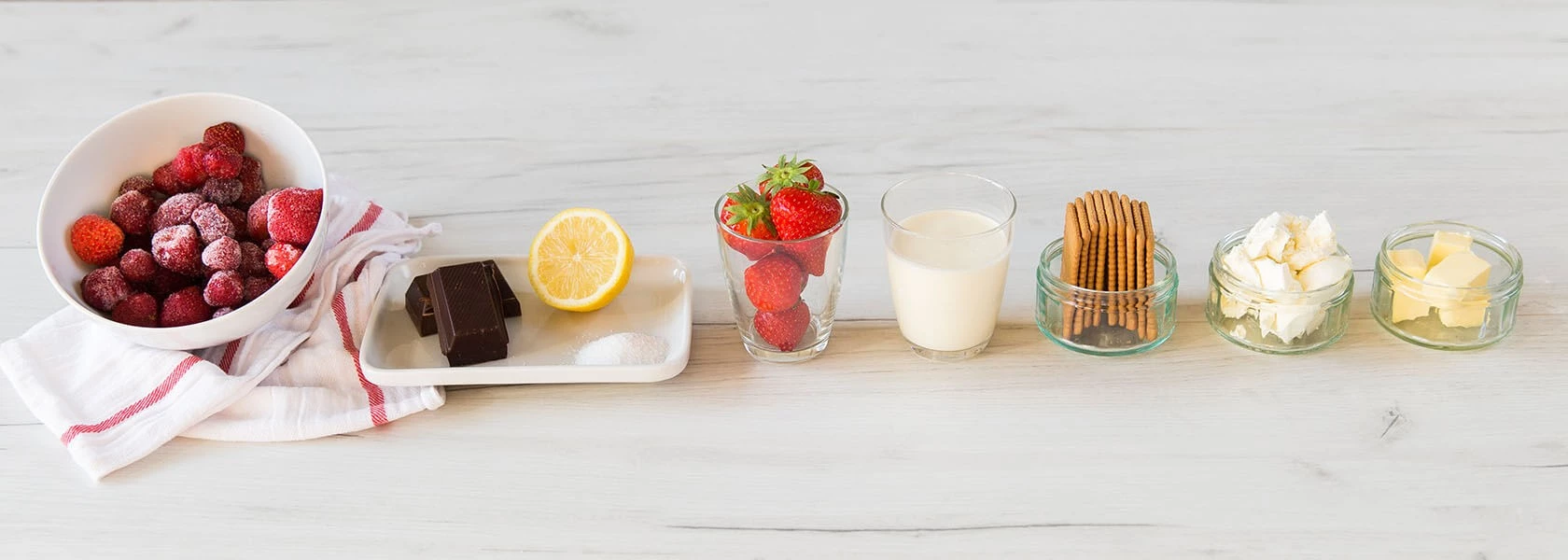 Verschiedene Dessertzutaten, darunter Erdbeeren, Schokolade, Zitrone, Zucker, Kekse, Milch, Schlagsahne, Butter.