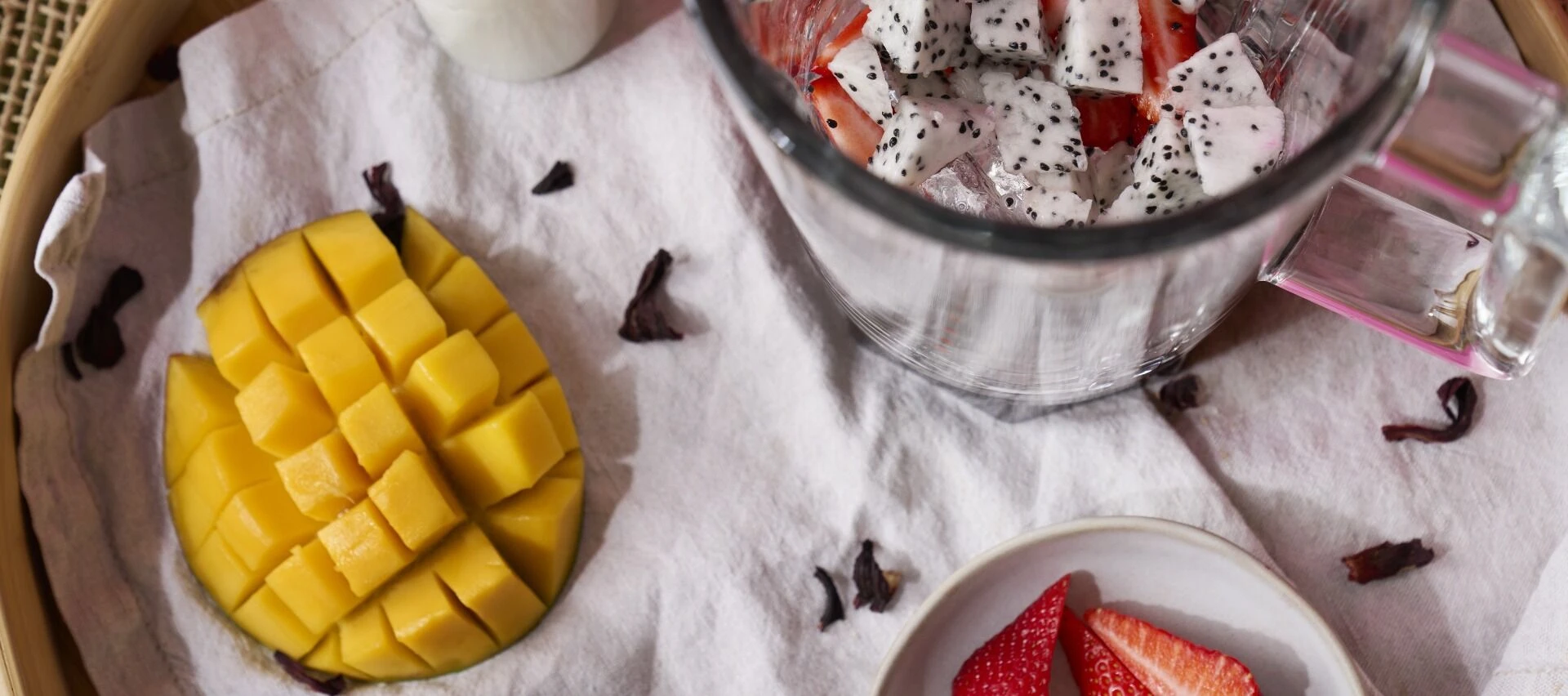 Ein Tablett mit gewürfelter Mango, eine Schale mit geschnittenen Erdbeeren und asiatischer Drachenfrucht, dazu ein Glas mit Stücken asiatischer Drachenfrucht und Erdbeeren - alles kunstvoll auf weißem Tuch angerichtet, ideal zum Genießen mit Früchtetee.