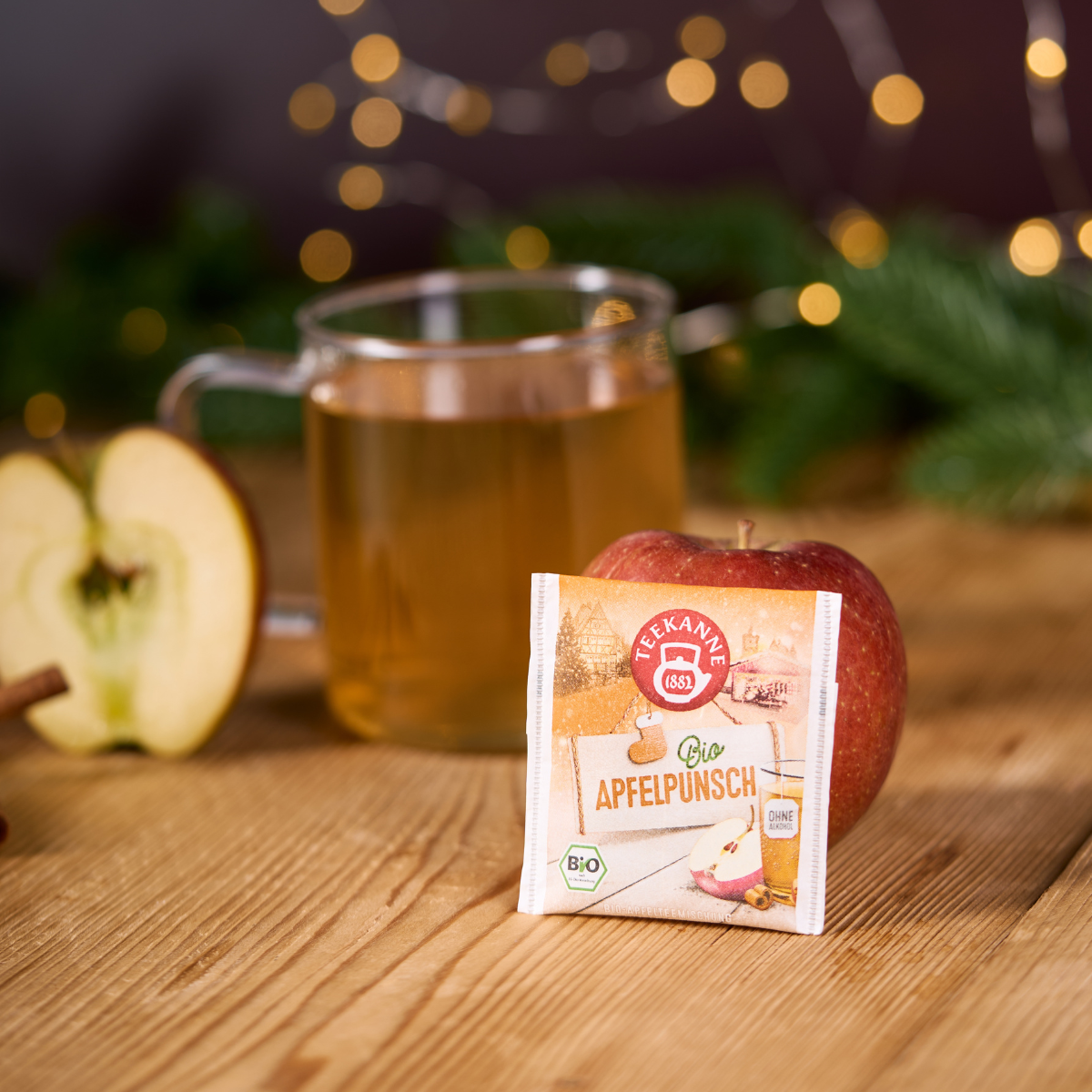 Eine Packung Bio Apfelpunsch liegt auf einem Apfel, im Hintergrund stehen ein Glas Apfelpunsch, ein halber Apfel, eine Zimtstange und festliche Lichter auf einem Holztisch.