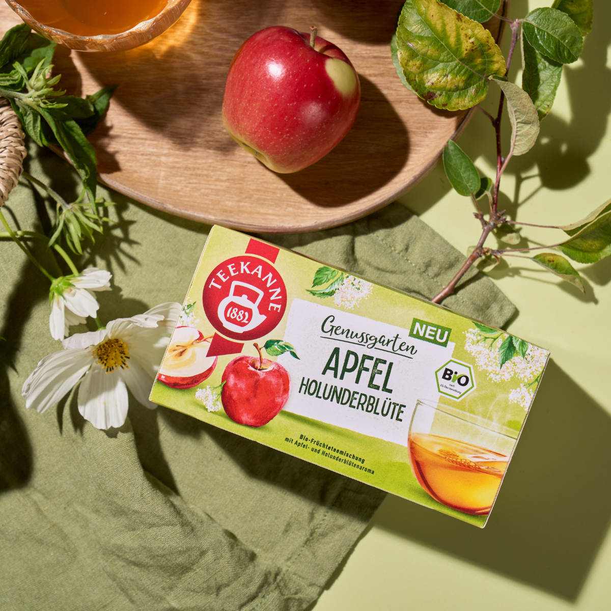 Eine Schachtel Bio Apfel Holunderblüte Tee liegt auf einem grünen Tuch neben einem frischen Apfel, weißen Blüten und grünen Blättern, alles in helles Tageslicht getaucht.