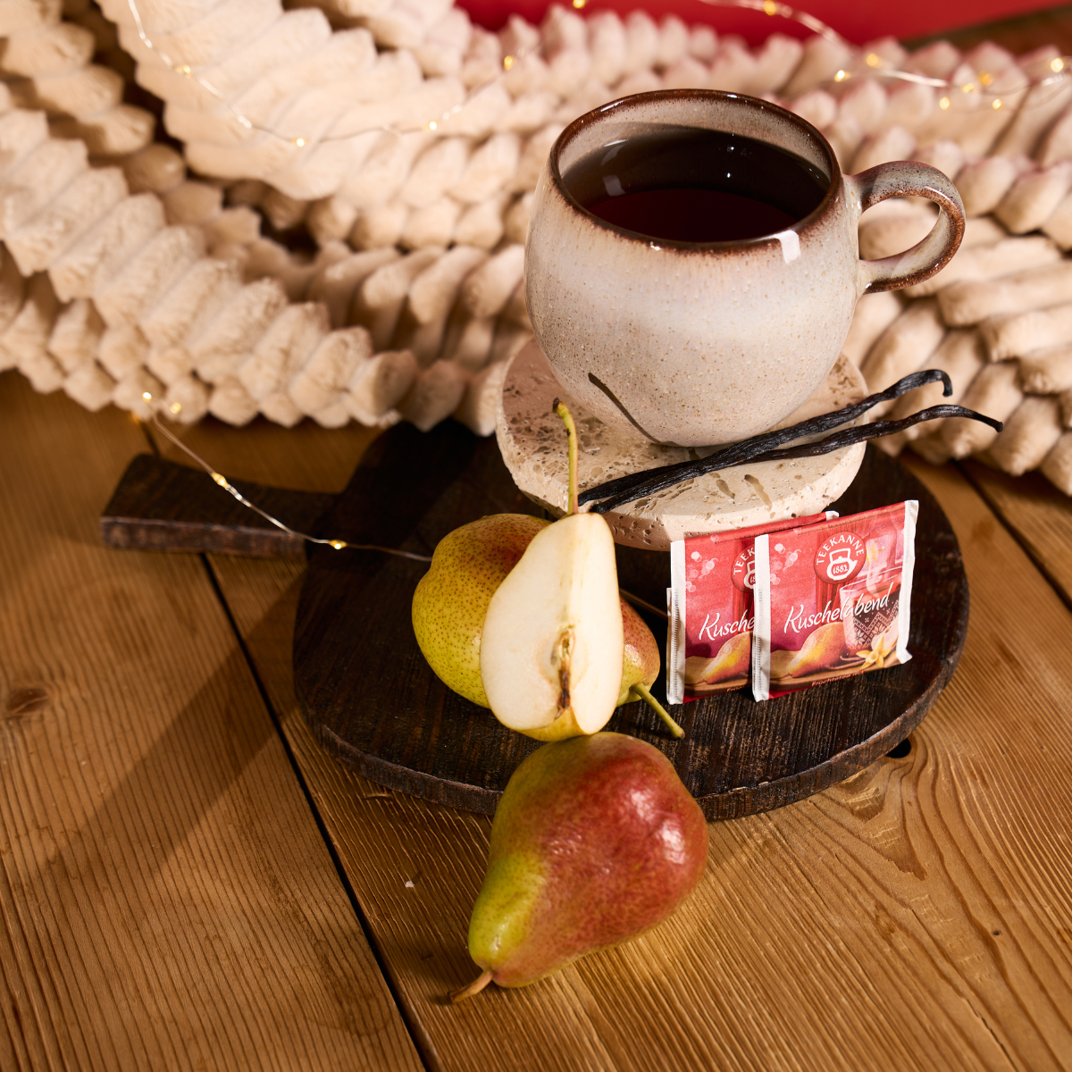 Der Kuschelabend-Keramikbecher steht auf einem Holzbrett mit Teebeuteln, geschnittener und ganzer Birne und Vanillebohnen - ideal für gemütliche Weihnachtsstimmung. Sanft leuchtende Lichterketten und Papiergirlanden sorgen für eine festliche Kulisse.