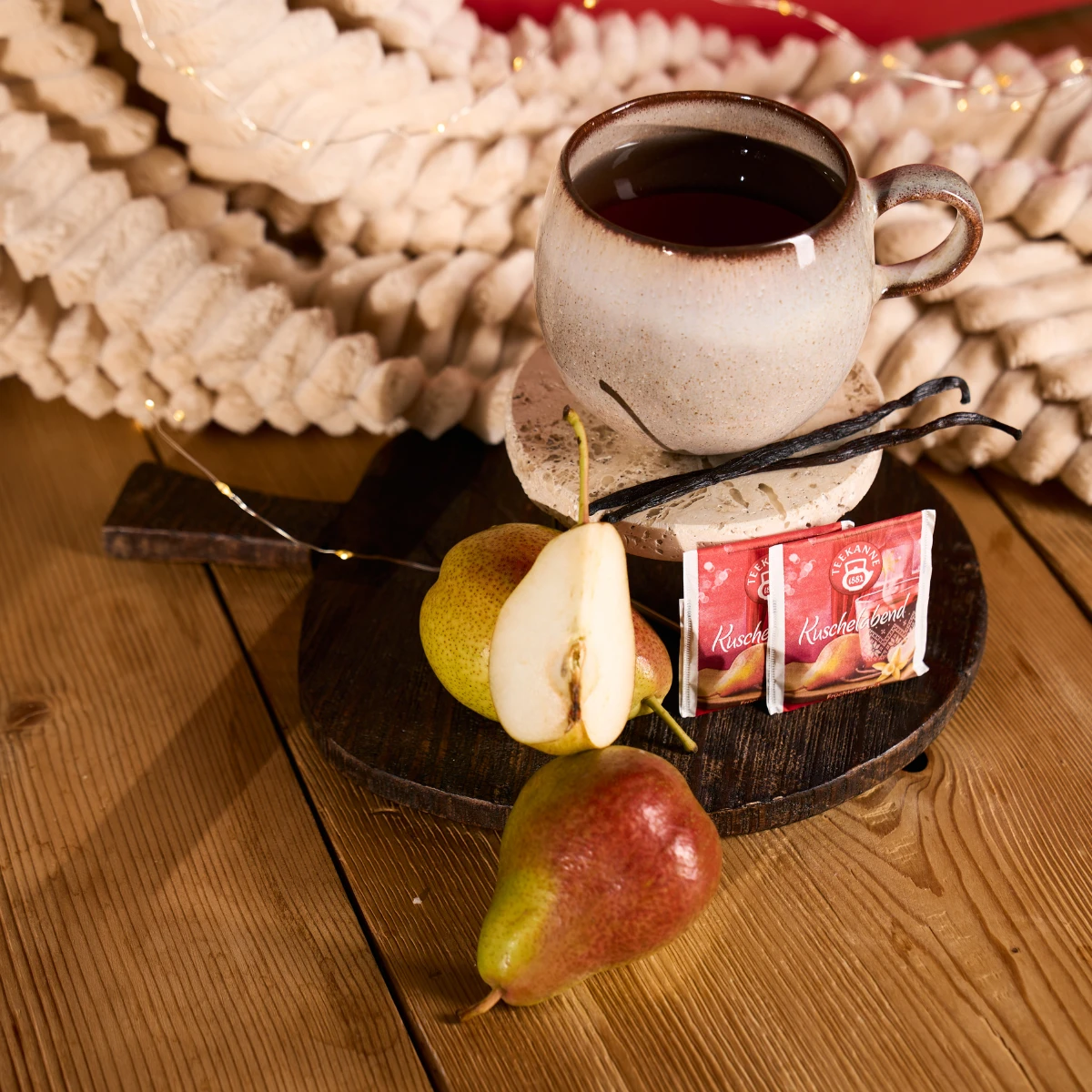 Der Kuschelabend-Keramikbecher steht auf einem Holzbrett mit Teebeuteln, geschnittener und ganzer Birne und Vanillebohnen - ideal für gemütliche Weihnachtsstimmung. Sanft leuchtende Lichterketten und Papiergirlanden sorgen für eine festliche Kulisse.