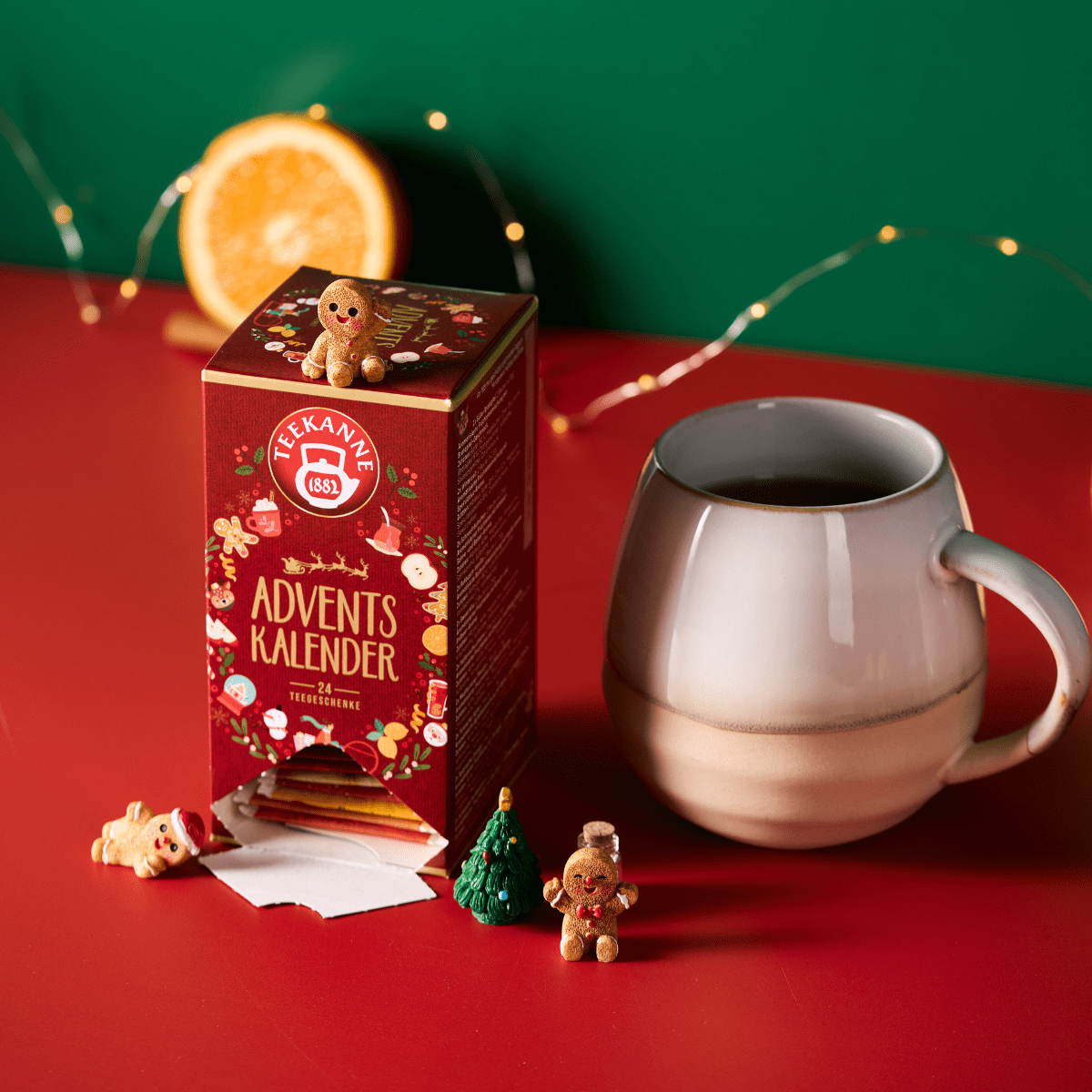 Eine festliche Schachtel mit Tee-Adventskalender steht auf einer roten Fläche mit Minifiguren, einem kleinen Baum, einer halben Orange, einem grauen Becher für den Teegenuss und einer Lichterkette - alles vor einem grünen Hintergrund.