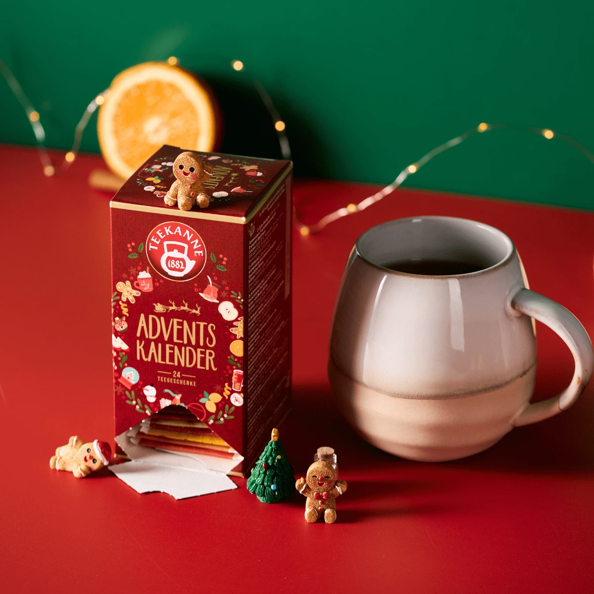 Eine festliche Schachtel mit Tee-Adventskalender steht auf einer roten Fläche mit Minifiguren, einem kleinen Baum, einer halben Orange, einem grauen Becher für den Teegenuss und einer Lichterkette - alles vor einem grünen Hintergrund.
