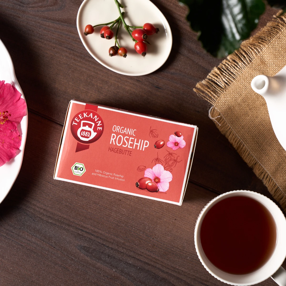 Eine Schachtel Bio-Hagebuttentee steht auf einem Tisch mit einer Tasse Tee, einer weißen Teekanne, Hibiskusblüten, Hagebutten in einer Schale und grünen Blättern - perfekt für alle, die einen Bio-Kräutertee kaufen möchten.