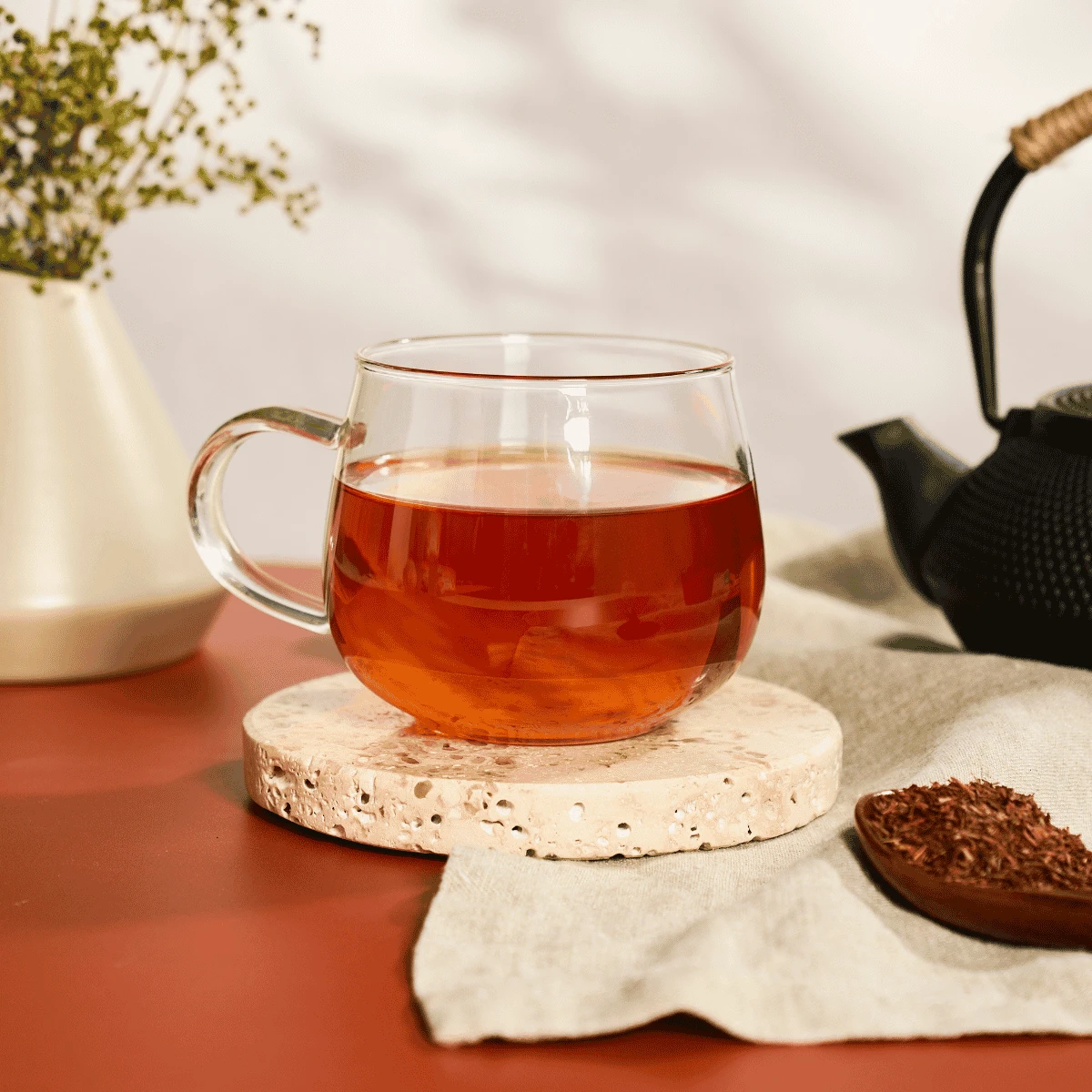 Ein Glas mit Bio-Rooibos aus Südafrika steht auf einem runden Untersetzer aus Stein, neben einer schwarzen Teekanne, einer weißen Vase mit Grünpflanzen und einem Löffel losem Tee auf einem beigen Tuch – alles auf einem roten Tisch angeordnet.