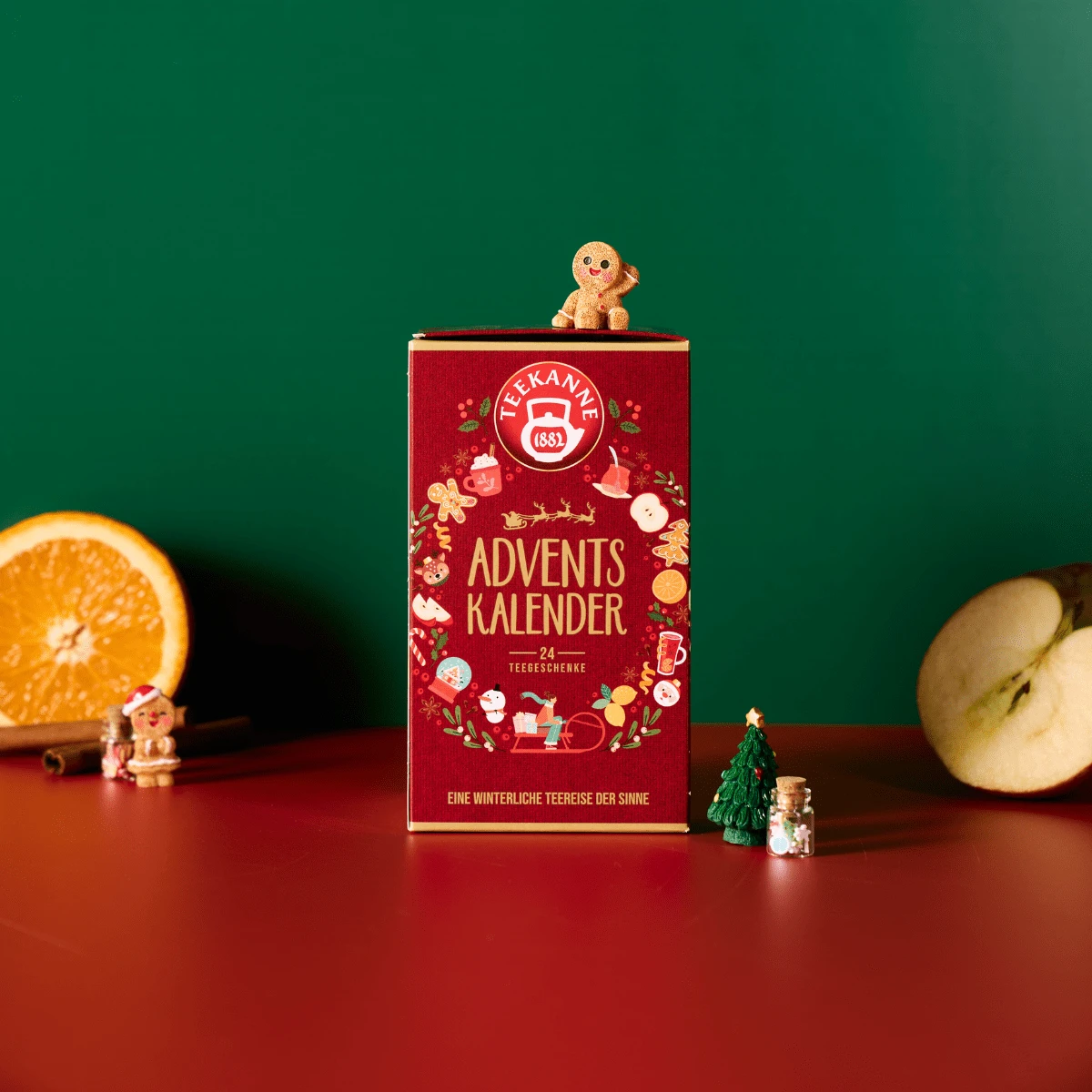 Eine festliche rote Schachtel mit Tee Adventskalender steht auf einer roten Fläche mit Orangen- und Apfelscheiben, Zimtstangen, einer Lebkuchenfigur, kleinen Figürchen und einem grünen Hintergrund - perfekt für Weihnachten und den Winter.