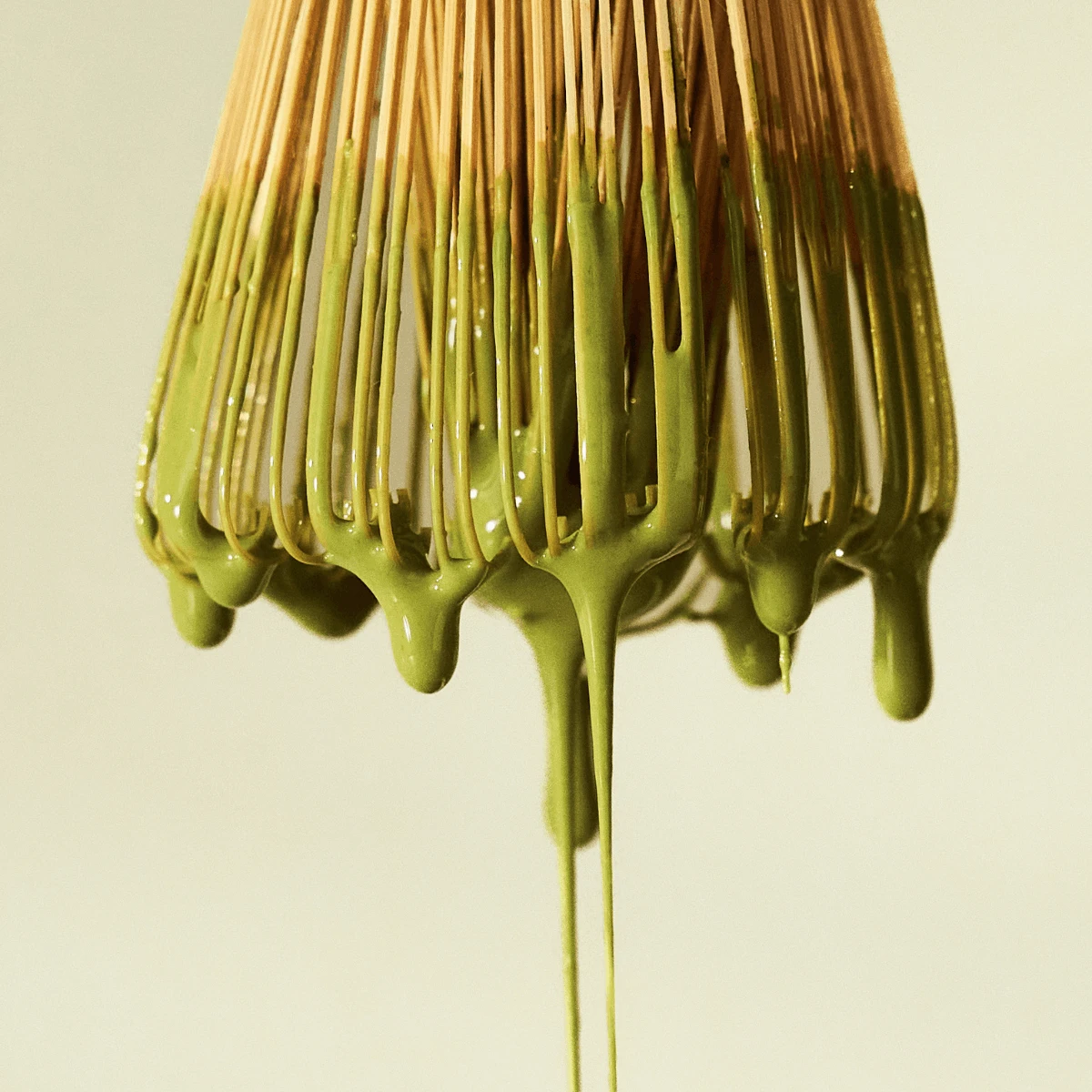 Ein Bio-Matcha-Bambusbesen, der mit leuchtend grünem Tee überzogen ist, wobei Matcha sanft von seinen Borsten auf einen hellen Hintergrund tropft.