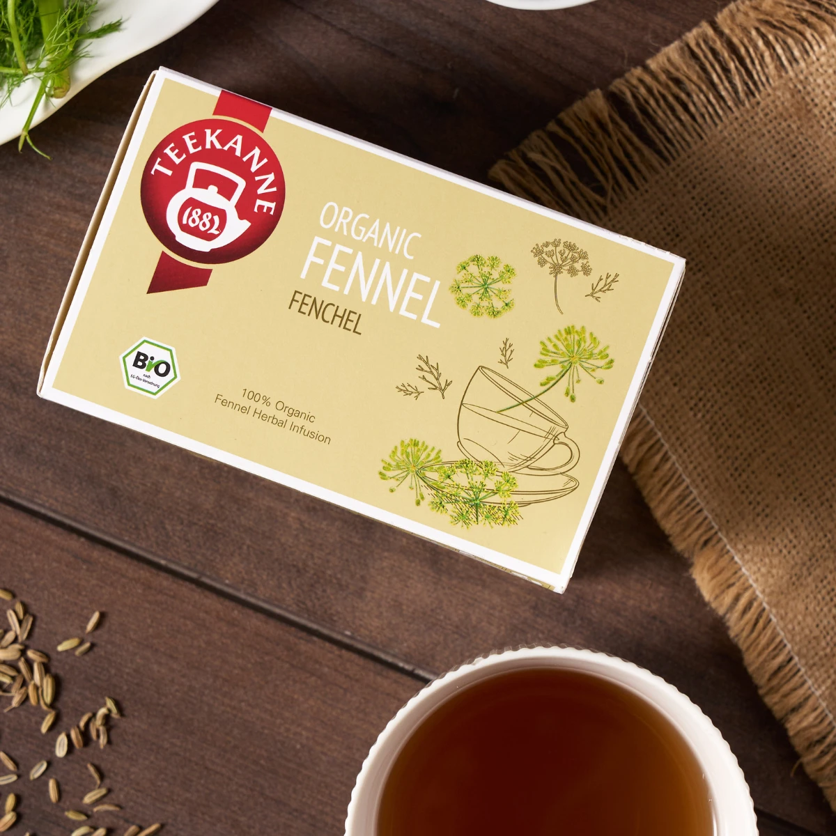 Eine Schachtel Bio-Fencheltee steht auf einem Tisch neben einer Tasse mit aufgebrühtem Tee, losen Fenchelsamen und frischem Fenchel. Die Schachtel zeigt ein grünes Fenchelmotiv und eine Teetasse - ideal für Kräutertee-Fans, die Bio-Tee kaufen möchten.