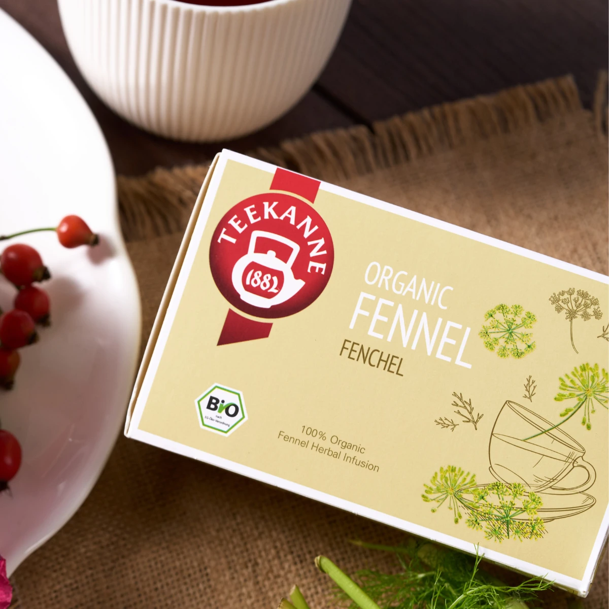 Eine Schachtel Bio-Fencheltee liegt auf einer braunen Matte neben einem weißen Teller mit roten Beeren und einer Tasse. Die Verpackung zeigt ein grünes Fenchelmotiv und eine Bio-Zertifizierung - ideal für alle, die Bio-Tee kaufen möchten.