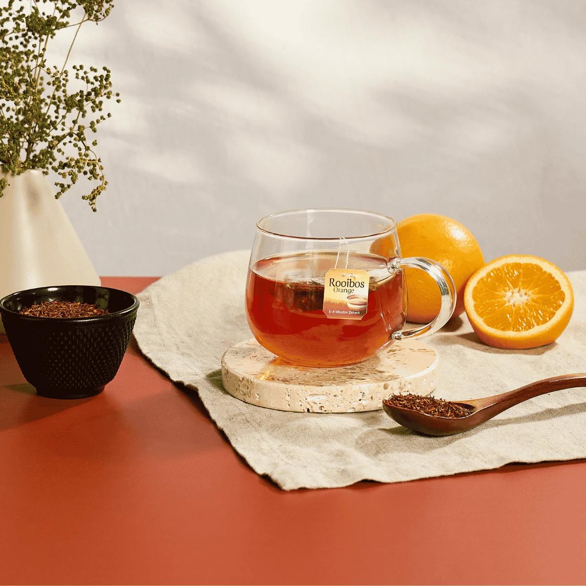 Auf einem roten Tisch mit beigem Tischtuch steht ein Glasbecher mit „Bio Rooibos Orange mild“, einem milden, fruchtigen Bio-Tee, auf einem Untersetzer neben einer halbierten Orange, einer schwarzen Schale mit Teeblättern, einem Holzlöffel mit Tee und eine
