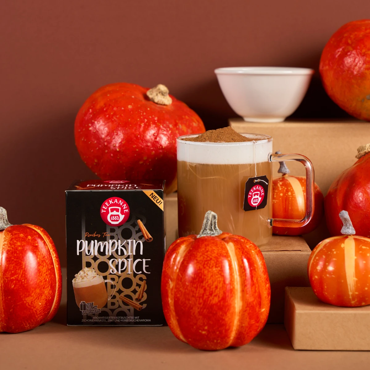 Ein Glasbecher Pumpkin Spice steht neben einer Schachtel Teekanne Pumpkin Spice Tee und Rotbusch, umgeben von Minikürbissen, einer Schale und braunen Schachteln auf einem warmen braunen Hintergrund für gemütliche Herbststimmung.