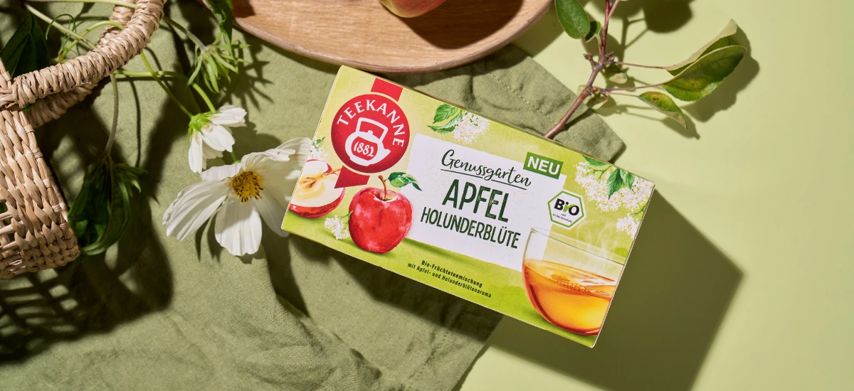 Eine Schachtel Teekanne Bio Apfel Holunderblüte liegt auf einem grünen Tuch neben einer weißen Blume, einem Apfel und einem Weidenkorb. Auf der Verpackung sind Äpfel und Holunderblüten abgebildet.