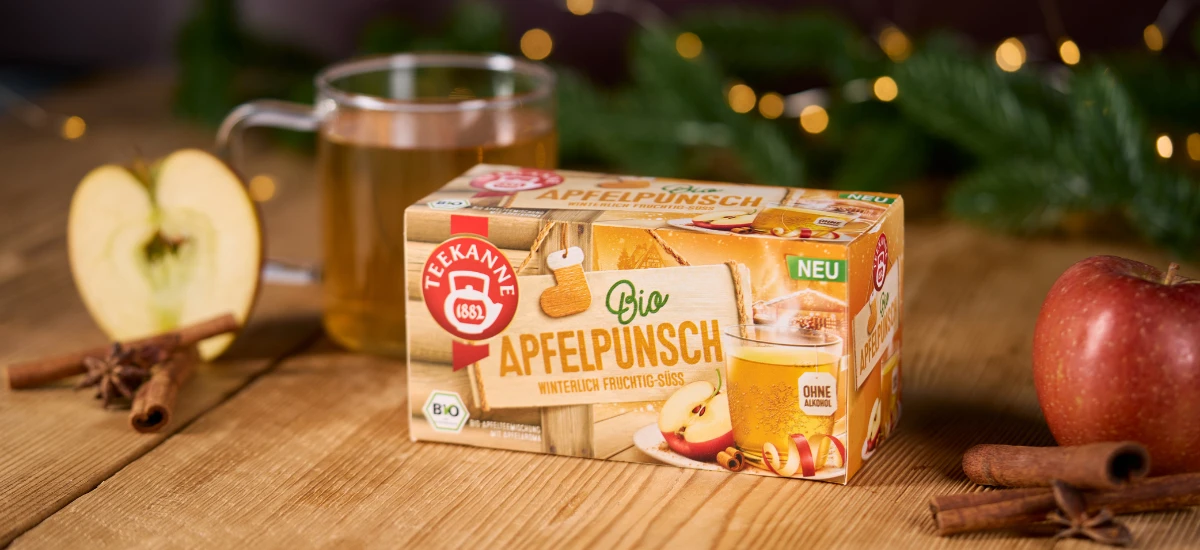 Eine Kiste Bio Apfelpunsch-Tee steht auf einem Holztisch, umgeben von Äpfeln, Zimtstangen, Sternanis, einem Glas Apfelpunsch und festlichem Grünzeug. Warme Lichter im Hintergrund schaffen eine gemütliche und einladende Atmosphäre.