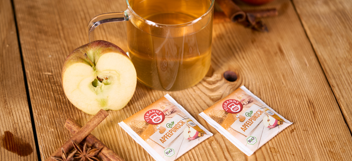 Auf einem Holztisch steht eine Glastasse mit Apfeltee, daneben ein aufgeschnittener Apfel, Zimtstangen, Sternanis und zwei Päckchen Bio-Apfelpunsch mit Apfelbildern - perfekt für einen wärmenden Bio-Wintertee.