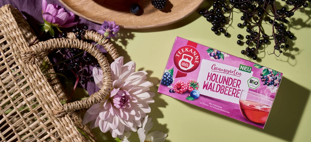 Eine Schachtel Bio Holunder Waldbeere Tee ruht auf einer hellgrünen Fläche, um die herum frische Holunderbeeren, Brombeeren, Blumen und ein geflochtener Korb für eine fruchtige, einladende Atmosphäre sorgen.