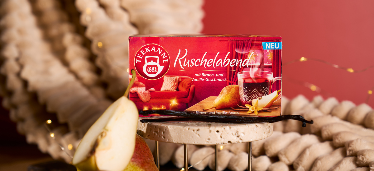 Eine Schachtel Kuschelabend-Tee steht auf einem Ständer neben einer frischen Birnenscheibe und einer Vanilleschote. Die gemütliche Dekoration und die warme Beleuchtung im Hintergrund vermitteln weihnachtliche Gemütlichkeit.