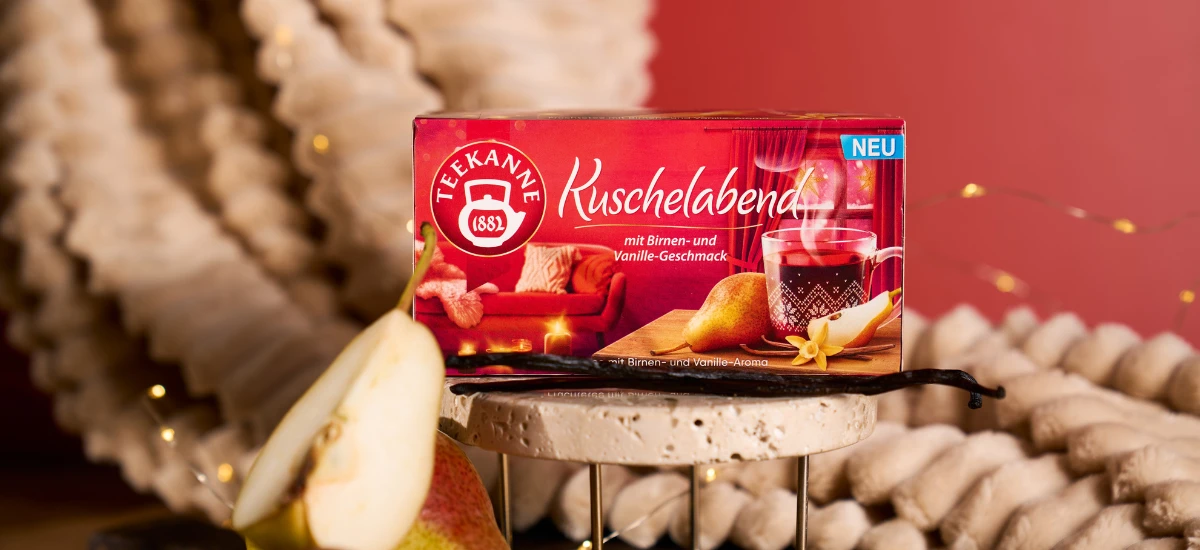 Eine Schachtel Kuschelabend-Tee steht auf einem Ständer neben einer frischen Birnenscheibe und einer Vanilleschote. Die gemütliche Dekoration und die warme Beleuchtung im Hintergrund vermitteln weihnachtliche Gemütlichkeit.