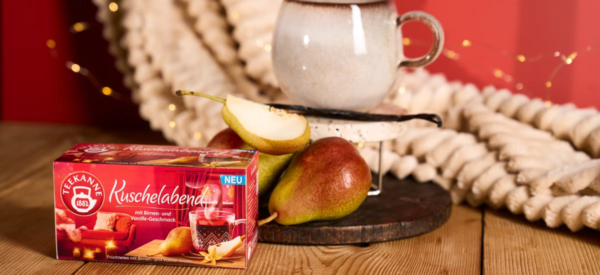 Eine Kiste Kuschelabend-Tee steht auf einem Holztisch, daneben zwei Birnen, eine Vanilleschote und eine Tasse. Eine weiche Strickdecke und Lichterketten bilden die perfekte Kulisse für diesen gemütlichen Früchtetee-Moment.