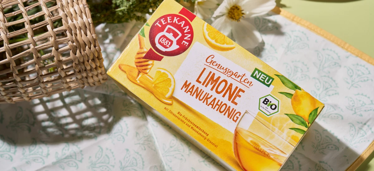Eine Schachtel Bio Limone Manukahonig Tee steht auf einem Tuch neben einer Korbdekoration und einer weißen Blume. Auf der gelben Verpackung sind Limone und Honig abgebildet, was den frischen, süßen Geschmack des Tees unterstreicht.