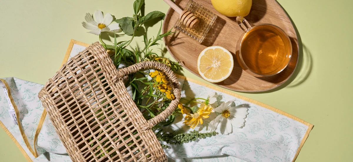Ein Weidenkorb mit gelben und weißen Blumen steht neben einem Tablett mit Tee, Bio-Zitronen und Bio-Limone Manukahonig auf einer hellgrünen Fläche und lädt zu einem Genussgarten-Moment ein.