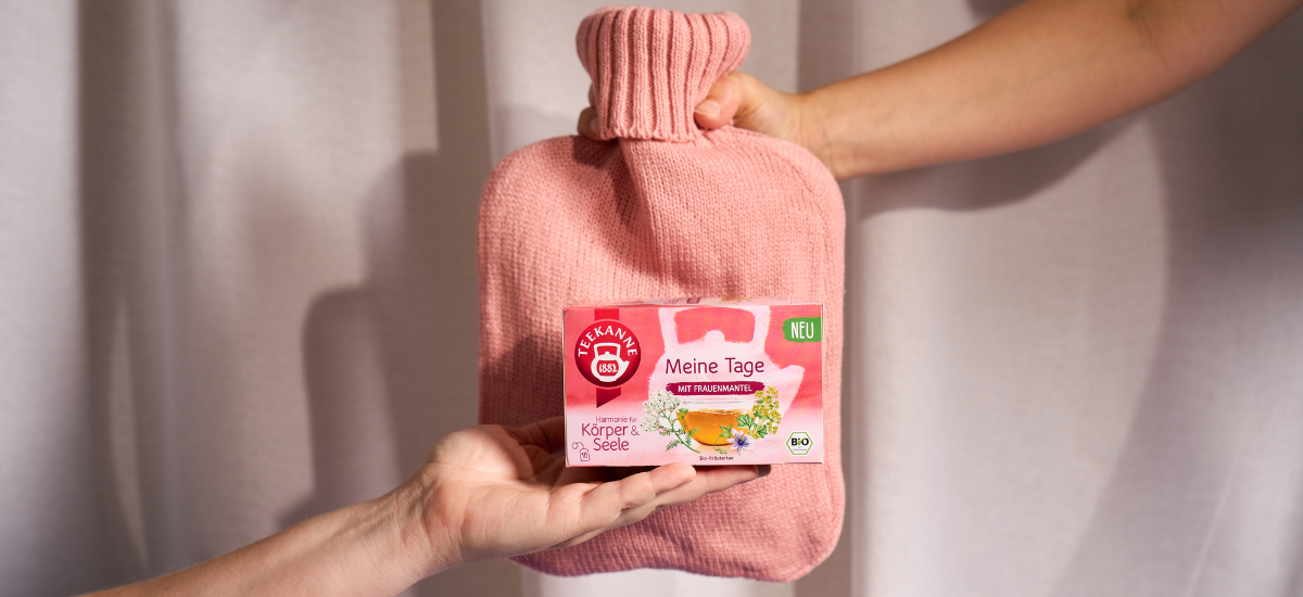 Zwei Hände halten eine rosafarbene Wärmflasche und eine Schachtel mit Meine Tage Tee mit Kräutern wie Salbei und Frauenmantel. Die Schachtel, die vor einem hellen Vorhang abgebildet ist, zeigt eine Teetasse, Blumen und ein grünes "NEU"-Etikett.