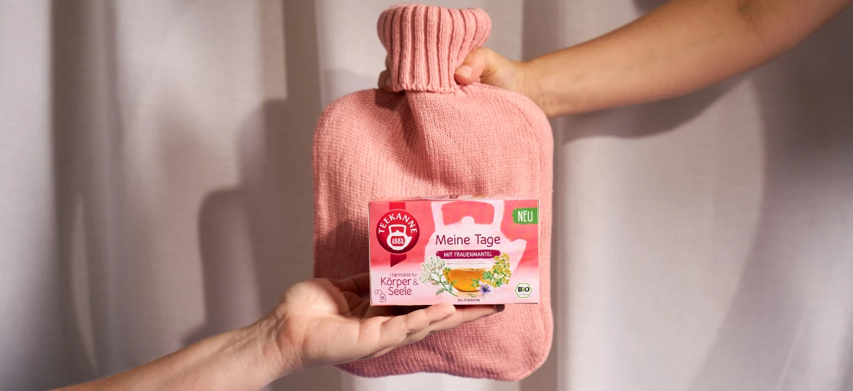 Zwei Hände halten eine rosafarbene Wärmflasche und eine Schachtel mit Meine Tage Tee mit Kräutern wie Salbei und Frauenmantel. Die Schachtel, die vor einem hellen Vorhang abgebildet ist, zeigt eine Teetasse, Blumen und ein grünes "NEU"-Etikett.