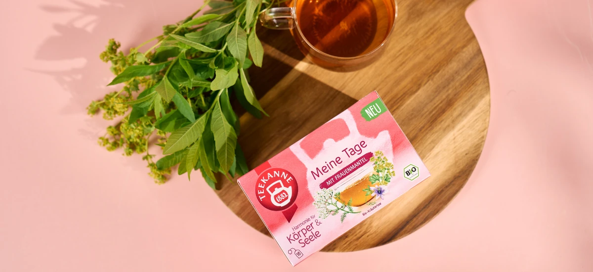 Eine Schachtel Kräutertee "Meine Tage" steht auf einem Holzbrett neben einer Glastasse mit Tee und frischen grünen Kräutern auf einem rosa Hintergrund.
