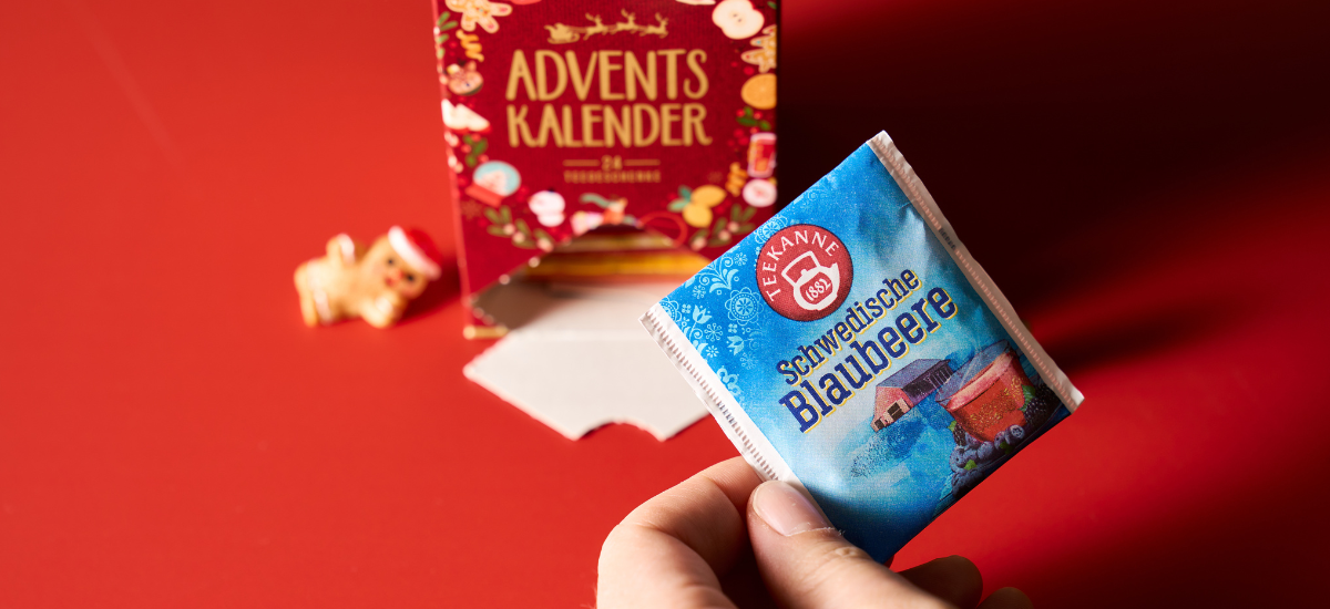 Eine Hand hält ein Päckchen Tee-Adventskalender-Tee vor einer roten Adventskalender-Box mit einem kleinen dekorativen Schwein auf der linken Seite, alles ruht auf einer roten Oberfläche für ein gemütliches Wintergefühl.