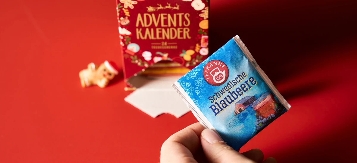 Eine Hand hält ein Päckchen Tee-Adventskalender-Tee vor einer roten Adventskalender-Box mit einem kleinen dekorativen Schwein auf der linken Seite, alles ruht auf einer roten Oberfläche für ein gemütliches Wintergefühl.