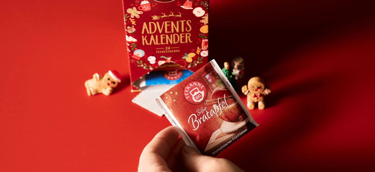 Eine Hand hält ein Päckchen mit der Aufschrift Tee Adventskalender vor einer festlichen roten Schachtel mit kleinen Lebkuchenfiguren und einem roten Hintergrund, der an die gemütliche Stimmung von Weihnachten und Winter erinnert.
