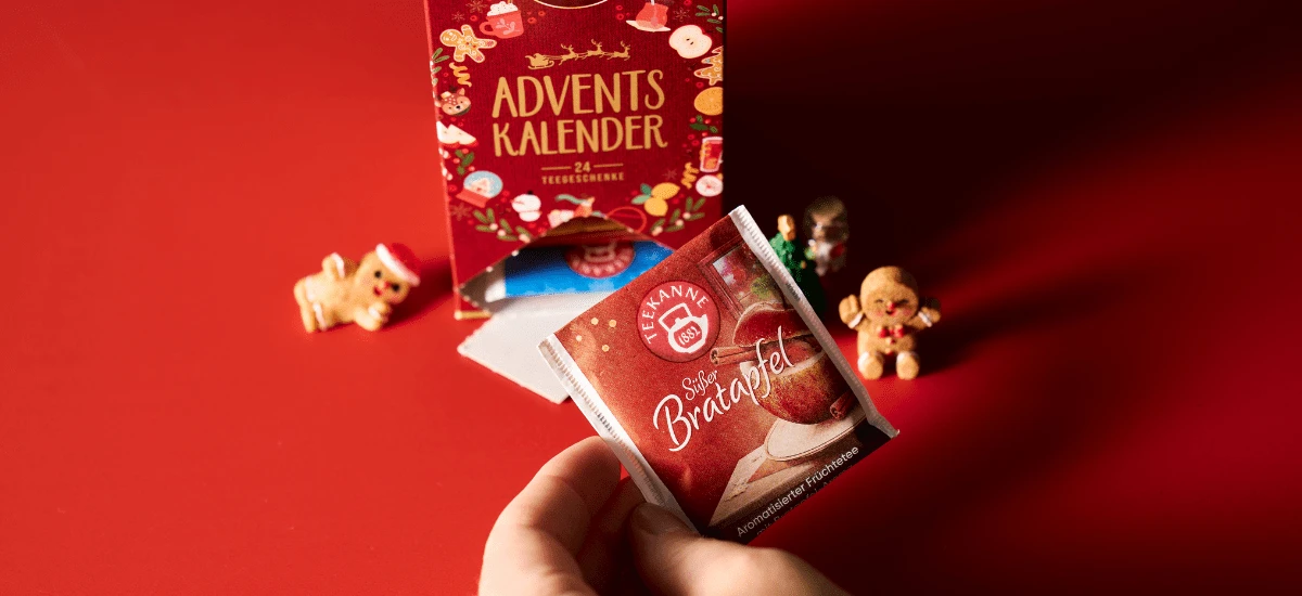 Eine Hand hält ein Päckchen mit der Aufschrift Tee Adventskalender vor einer festlichen roten Schachtel mit kleinen Lebkuchenfiguren und einem roten Hintergrund, der an die gemütliche Stimmung von Weihnachten und Winter erinnert.