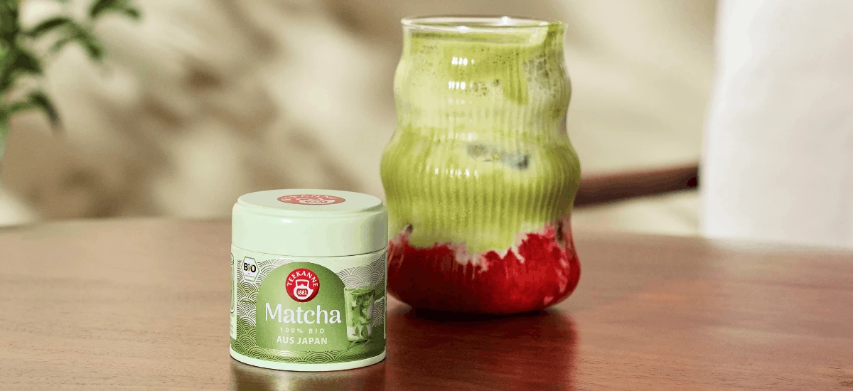 Auf einem Holztisch steht eine kleine Dose Bio-Matcha neben einem Glas mit einem Getränk, das grüne und rote Schichten aufweist – wahrscheinlich ein Erdbeer-Matcha-Latte.