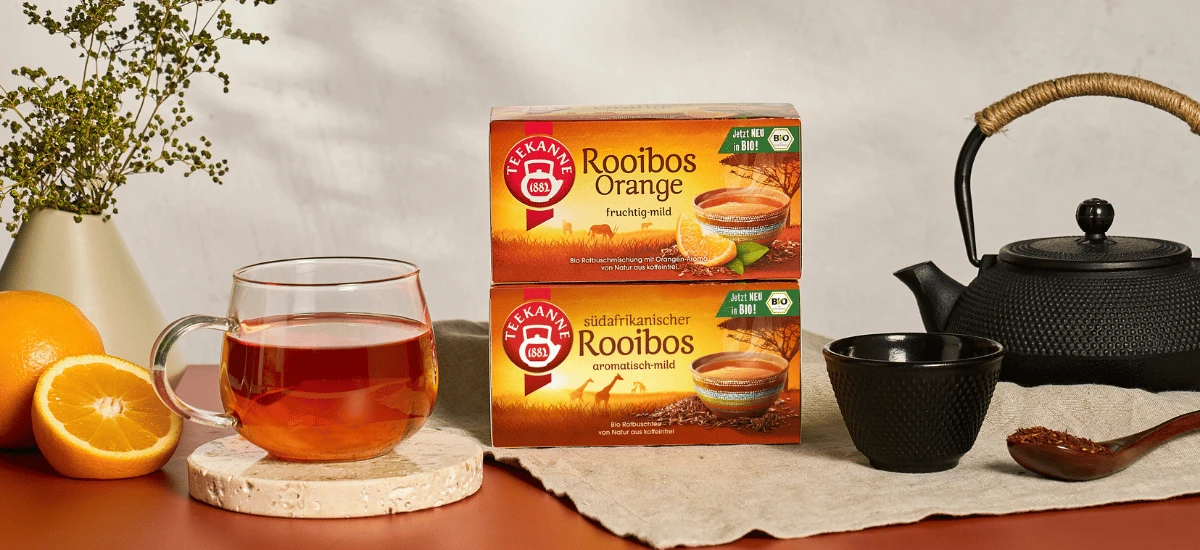 Auf einem Untersetzer steht eine Glastasse mit mildem, fruchtigem Bio-Rooibos-Orangen-Tee neben zwei Schachteln mit Bio-Rooibos-Orangen-Tee, einer Orange, einer schwarzen Teekanne, einer schwarzen Tasse, losen Teeblättern und einer mit Grünpflanzen gefüll