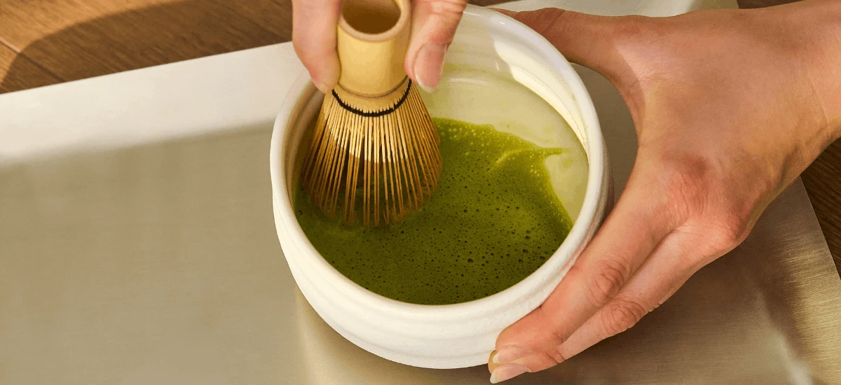 Eine Person verrührt mit einem Bambusbesen Bio-Matcha in einer weißen Schale zu einem schaumigen, leuchtend grünen Tee.
