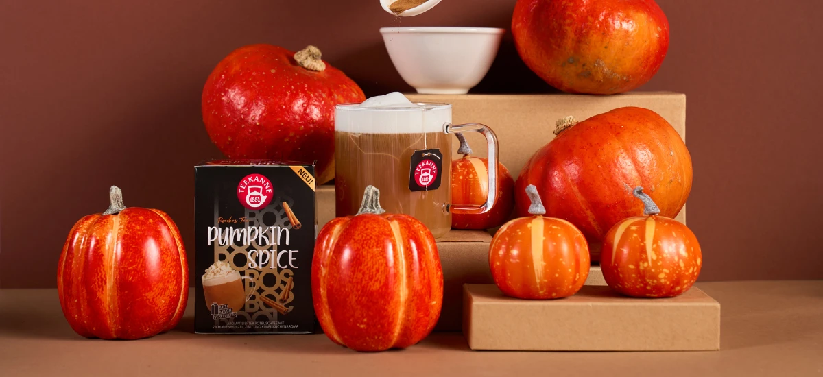 Ein Glasbecher mit Pumpkin Spice Latte steht zwischen kleinen Kürbissen und einer Schachtel mit der Aufschrift Pumpkin Spice auf braunen Kartons - perfekt für gemütliche Herbstmomente und den Genuss von Kürbiskuchen.