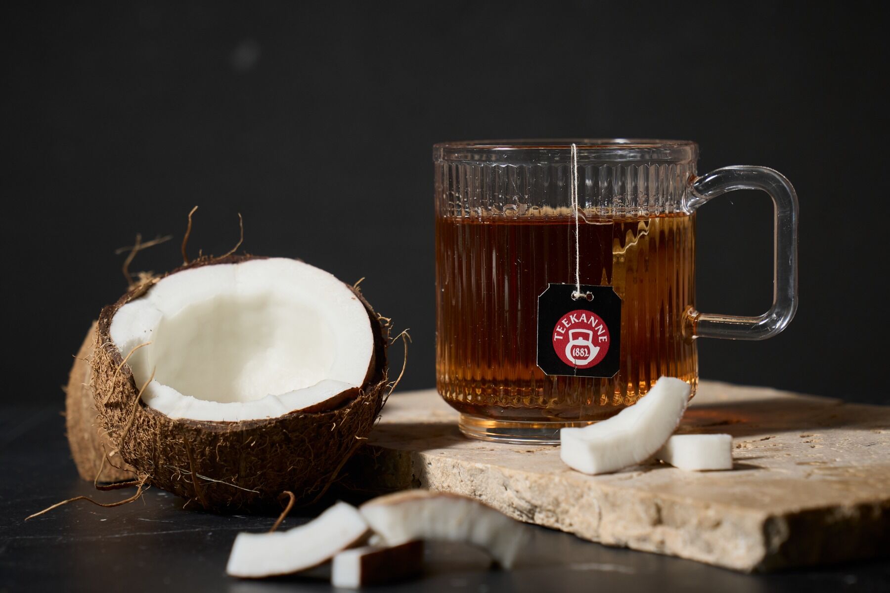 Eine Glastasse mit schwarzem Tee „Coco & Cream“ steht auf einer Steinplatte neben einer halbierten Kokosnuss und Kokosnussstücken vor einem dunklen Hintergrund.