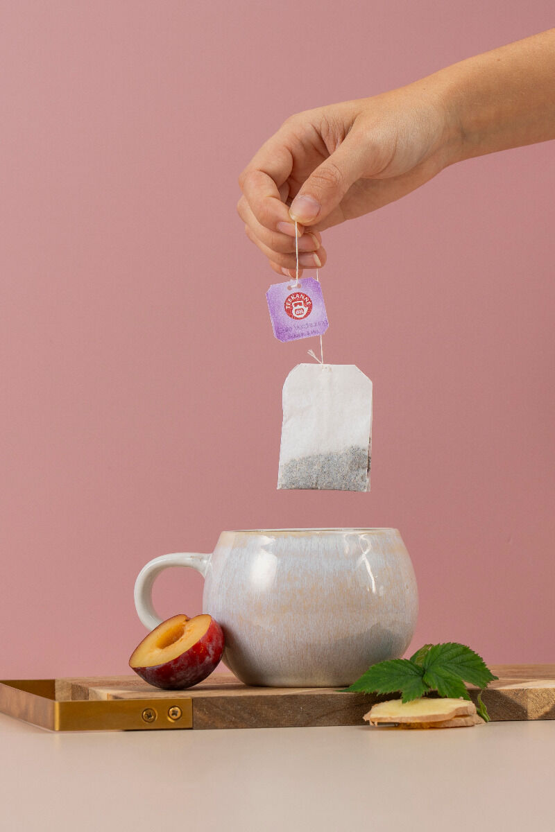 Eine Hand taucht einen Teebeutel mit violettem Etikett in eine beige Tasse auf einem Holztablett, umgeben von Pflaume, Minze und Ingwer auf rosa Untergrund – eine perfekte Szene für Ihr Harmonie für Körper & Seele Mix & Match.
