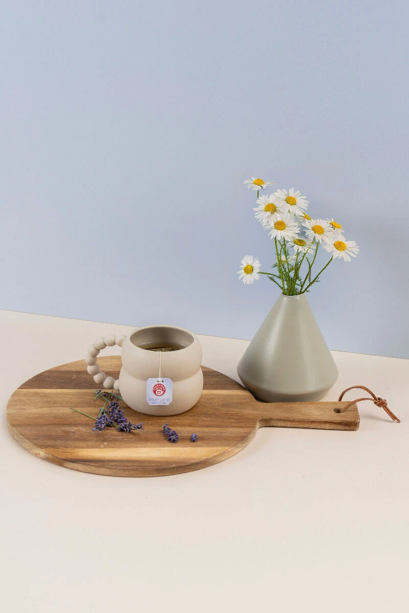 Eine beige Tasse mit Kräutertee und Teebeutel, auf einem runden Holztablett neben Lavendelzweigen und einer grauen Vase mit weißen Gänseblümchen, präsentiert den Harmonie für Körper & Seele Mix & Match vor hellem Hintergrund.