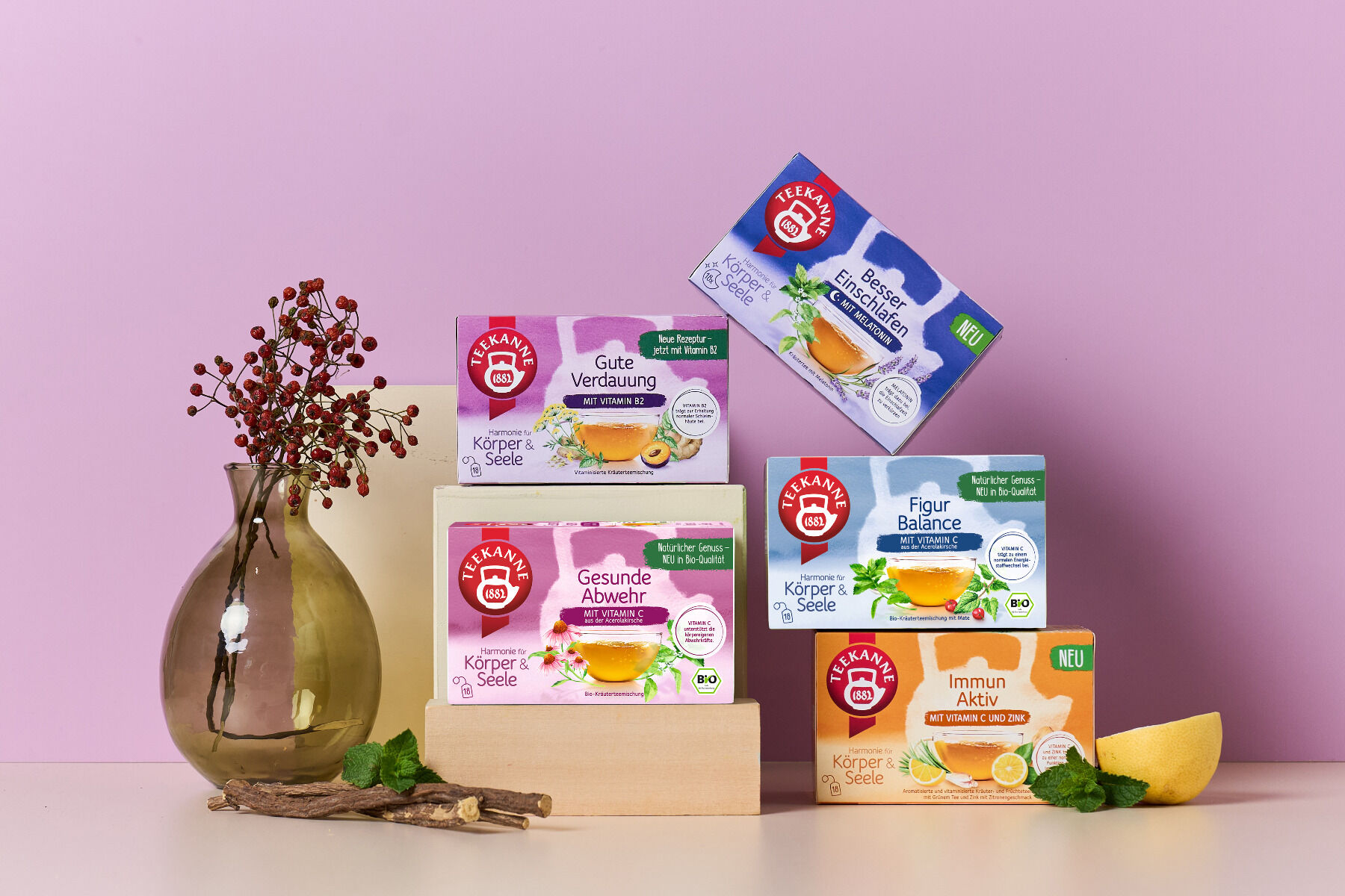 Fünf Schachteln „Harmonie für Körper & Seele Mix & Match Kräutertees“ mit deutschen Etiketten sind auf Holzklötzen vor einem hellvioletten Hintergrund angeordnet, neben einer Glasvase mit roten Beeren, Grünpflanzen und einer Zitronenscheibe in der Ecke.